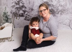 Lächelnde Frau mit Brille hält ein Kind auf dem Schoß vor einer winterlichen Fotokulisse
