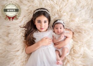 Kind und Baby in weißen Kleidern liegen nebeneinander auf einer hellen Decke, mit AFNS-Award-Winner-Siegel Juli 2024