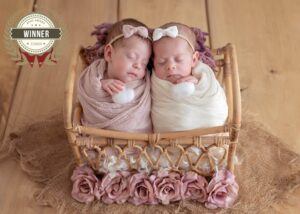 Zwei schlafende Babys mit Schleifen im Haar liegen in einem Korbbett mit Rosen, mit AFNS-Award-Winner-Siegel Juli 2024