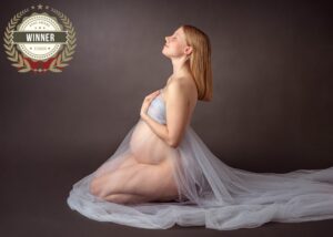 Schwangere Frau kniet im Profil mit grauem Tüll vor dunklem Hintergrund, mit AFNS-Award-Winner-Siegel Juli 2024