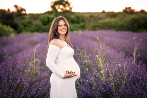 Schwangere Frau im weißen Kleid steht zwischen blühendem Lavendel und hält den Schriftzug Liebe vor ihren Bauch