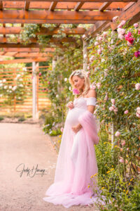 Schwangere Frau im langen rosa Kleid steht unter einer Pergola neben blühenden Rosen