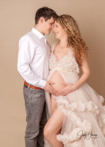 Ein Paar berührt sich mit der Stirn und hält gemeinsam den Babybauch vor beigem Studiohintergrund