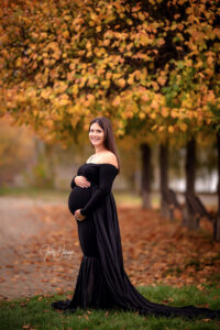 Schwangere Frau im langen schwarzen Kleid steht unter einem Baum mit goldgelbem Herbstlaub