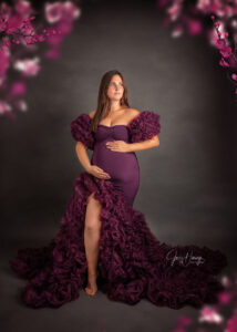 Schwangere Frau im violetten Kleid mit langer Rüschenschleppe vor dunklem Hintergrund und violetten Blüten