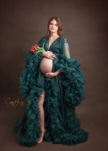 Schwangere Frau im dunkelgrünen Rüschenkleid hält ihren freien Babybauch vor braunem Studiohintergrund