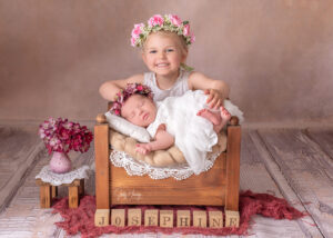 Kind mit Blumenkranz hält ein schlafendes Baby in einer kleinen Holzkiste