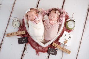 Zwei schlafende Babys liegen nebeneinander in einer herzförmigen Schale zwischen Uhren und Namenswürfeln