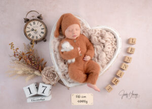 Schlafendes Baby in braunem Strickanzug in einer herzförmigen Schale neben Uhr und Namenswürfeln