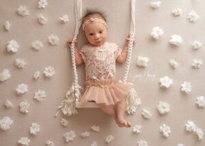 Baby in einem rosafarbenen Kleid auf einer kleinen Seilschaukel vor einer Wand mit weißen Blüten