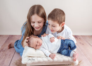 Zwei Kinder betrachten ein schlafendes Baby, das zwischen ihnen auf einem Kissen liegt