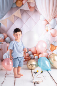 Barfüßiges Kind in blauem Hemd und Shorts steht vor pastellfarbenen Ballons und einer Wimpelkette