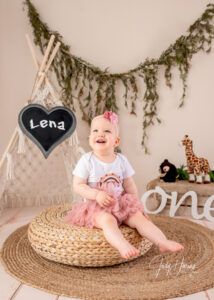 Lächelndes Kleinkind im rosa Rock sitzt vor einem kleinen Tipi mit Kreidetafel, Kuscheltieren und ONE-Schriftzug