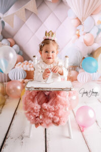 Kleinkind mit Krönchen und rosa Rüschenrock sitzt auf einem weißen Hochstuhl vor pastellfarbenen Ballons