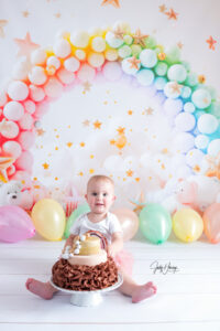 Kleinkind sitzt mit einer kleinen Torte vor einem regenbogenfarbenen Ballonbogen und Sternendekoration