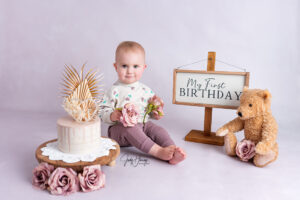 Kleinkind sitzt neben einer weißen Torte, einem Teddybären und einem Schild zum ersten Geburtstag
