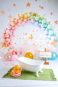 Kleine weiße Badewanne mit Badespielzeug auf grünem Teppich vor einer bunten Ballonkulisse